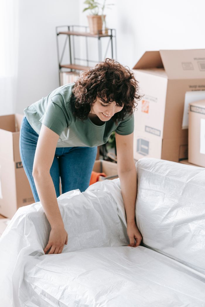 Woman wrapping a sofa with plastic for moving in a bright living room.